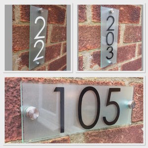 Könnte beinhalten: Rechteckige Hausnummernschilder aus Glas mit Edelstahlbeschlägen. Die Zahlen sind in einer fetten, modernen Schriftart gehalten. Der Hintergrund ist eine Backsteinmauer. Die Zahlen sind weiß und schwarz.