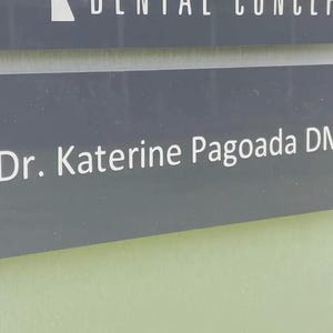 May include: A gray sign with the text "Dr. Katerine Pagoada DMD" in white lettering. The sign is mounted with two silver fasteners. The top of the sign reads "Dental Concepts" in white lettering.