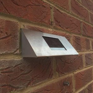 May include: A solar-powered outdoor light fixture mounted on a red brick wall. The light has a silver metal casing with a rectangular solar panel. The brick wall has a textured surface.