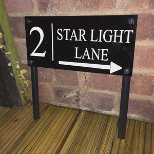 Könnte beinhalten: Schwarzes Straßenschild mit der Aufschrift "2 STAR LIGHT LANE" und einem weißen Pfeil nach rechts. Das Schild ist auf zwei schwarzen Pfosten montiert und steht vor einer Backsteinmauer.