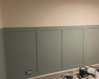 Puede incluir: Una habitación con un diseño de pared de dos tonos. La sección inferior está pintada de un verde apagado, dividida en paneles rectangulares. La pared superior es de color beige claro. Suministros de pintura visibles en el suelo.
