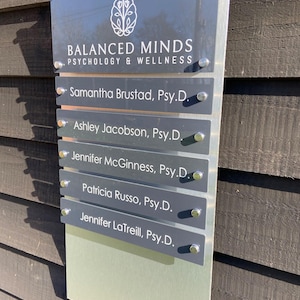 Puede incluir: Un letrero plateado con el texto "SUITE 319" y "BALANCED MINDS PSYCHOLOGY & WELLNESS". Debajo, placas con nombres individuales para los miembros del personal, incluyendo Samantha Brustad, Ashley Jacobson, Jennifer McGinness, Patricia Russo y Jennifer LaTreill.
