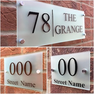 May include: Frosted glass house number signs with stainless steel standoffs. The top sign reads "78 THE GRANGE" in black lettering. The bottom signs display numbers and "Street Name". Mounted on a red brick wall.