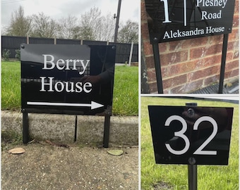May include: Black rectangular address signs with white lettering. One sign reads "Berry House" with an arrow. Another sign displays "1 Pleshey Road Aleksandra House". The third sign shows the number "32". All signs are mounted on black metal posts.