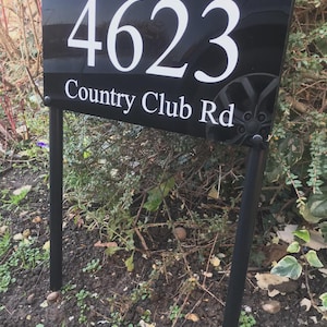May include: A black address sign displaying the number "4623" in white, with "Country Club Rd" beneath. The sign is supported by black metal posts and set against a backdrop of foliage.