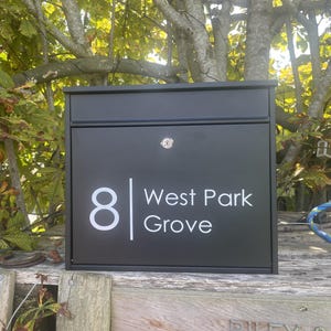 Könnte beinhalten: Ein schwarzer Briefkasten mit der Adresse "8 West Park Grove" in weißen Buchstaben. Der rechteckige Briefkasten hat ein Schloss und ist an einem Holzzaun befestigt, mit grünen Blättern und Ästen im Hintergrund.