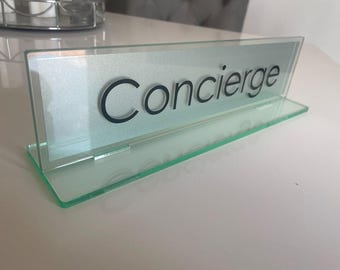 May include: A clear glass desk sign displaying the word "Concierge" in black. The sign is rectangular with a slanted top and a base. The background behind the text is a light silver colour. The sign is made of glass.