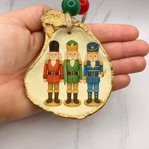 May include: A Christmas ornament featuring three toy soldier nutcrackers. The ornament is made from a gold-edged oyster shell and has a string for hanging. The nutcrackers are dressed in red, green, and blue uniforms.