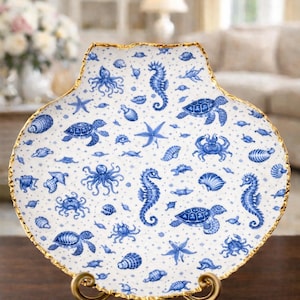 May include: A seashell-shaped decorative plate with a gold rim, featuring a blue and white nautical pattern of seahorses, turtles, starfish, and other sea creatures. The plate is displayed on a gold stand. The Etsy shop name is visible at the bottom.