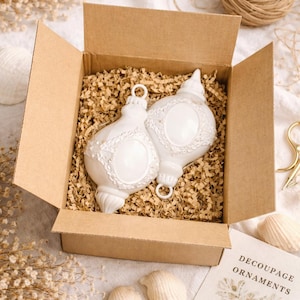 Könnte beinhalten: Zwei weiße, verzierte Decoupage-Ornamente mit silberfarbenen Metallaufhängern liegen in einer offenen Pappschachtel, die mit zerkleinertem Verpackungsmaterial gefüllt ist. Die Schachtel ist geöffnet und zeigt die Ornamente. Die Worte "DECOUPAGE ORNAMENTS" sind auf einer Karte gedruckt.