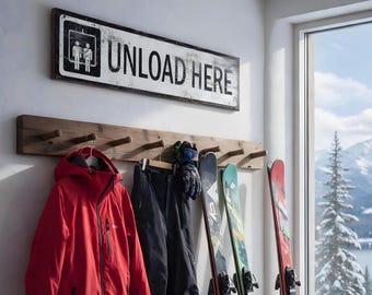 May include: A rectangular wall sign with a distressed white finish and black text reading UNLOAD HERE, featuring a graphic of two figures in an elevator. Below, a wooden coat rack holds a red hooded jacket, black pants, and gloves.
