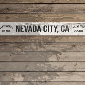May include: White wooden sign with black text that reads "San Francisco 165 Miles Nevada City, CA Elevation 2525 Feet". The sign is mounted on a wood plank wall.