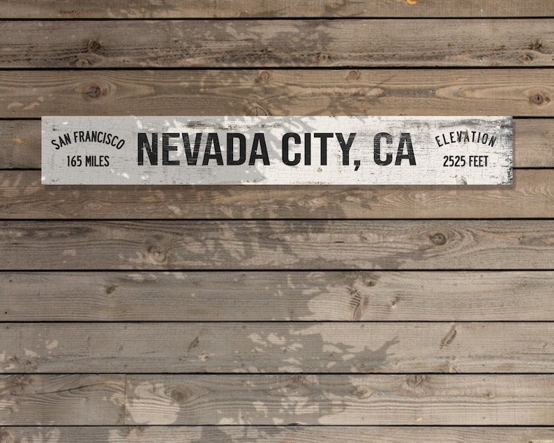May include: White wooden sign with black text that reads "San Francisco 165 Miles Nevada City, CA Elevation 2525 Feet". The sign is mounted on a wood plank wall.