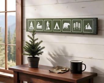 May include: A long, rectangular, dark green wooden wall sign featuring seven white graphic icons depicting a campfire, a person in a canoe, a hiker, a tent, a bear, pine trees, and a mountain peak, all enclosed in individual white-lined boxes.