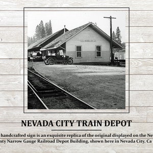 May include: Black and white photograph of a train depot building in Nevada City, California. The building has a sign that reads "Nevada City". A car is parked in front of the building.
