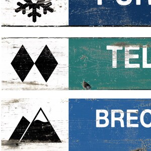 May include: A collage of wooden signs with a distressed finish. The signs feature white text and black graphic elements, including a snowflake, two black diamonds, and mountain silhouettes. The signs are painted in blue, green, and white.