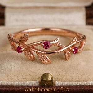 Può includere: Anello in oro rosa con un design a foglie e tralci, con tre piccole gemme rosse rotonde. L'anello è presentato in un portagioie marrone con interno beige. Il testo "Ankitgemcrafts" è visibile.