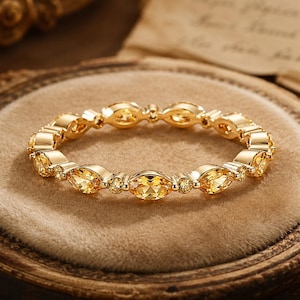 Puede incluir: Un anillo dorado con una banda continua de piedras preciosas amarillas, tanto ovaladas como redondas. El anillo se exhibe sobre una superficie de terciopelo beige dentro de un marco de madera decorativo. Las piedras están espaciadas uniformemente.