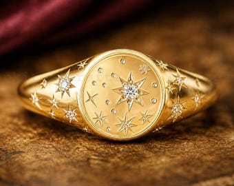 Puede incluir: Un anillo dorado con un diseño celestial. El anillo presenta un círculo central con una estrella grande y estrellas y puntos más pequeños. Estrellas adicionales están engastadas a lo largo de la banda. El anillo está sobre una superficie de madera.