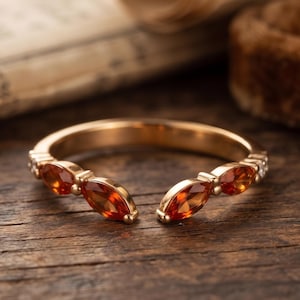Könnte beinhalten: Ein offener Ring aus warmfarbenem Metall mit vier orangefarbenen Edelsteinen im Marquise-Schliff, die paarweise auf beiden Seiten der Öffnung angeordnet sind. Das Band ist schmal und poliert und zeigt ein symmetrisches Design mit leuchtenden Steinen.