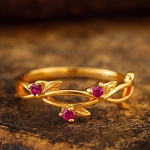 Puede incluir: Un anillo de oro con una banda diseñada con metal entrelazado en forma de enredadera y hojas. El anillo está engastado con tres pequeños rubíes redondos de color rojo rosado vibrante, situados entre los detalles de las hojas metálicas.
