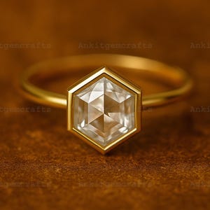 Puede incluir: Un anillo de oro con una gema clara hexagonal. El anillo tiene una banda delgada de oro. La gema está engastada en un bisel de oro. El fondo es de color marrón cálido. El texto "Ankitgemcrafts" se repite.