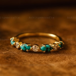 May include: A gold ring featuring alternating turquoise and clear gemstones. The ring's band is a polished gold, and the gemstones are set in a row around the band. The ring is displayed on a wooden surface.