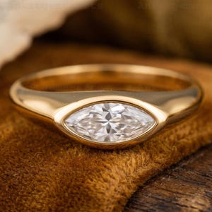 Op de afbeelding: Een gouden ring met een marquise-geslepen diamant. De diamant is gezet in een bezel-zetting en de band is glad en gepolijst. De ring wordt gepresenteerd op een bruin fluwelen oppervlak.