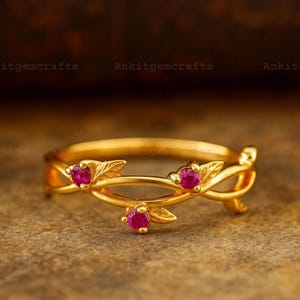 May include: A gold-colored ring with a floral design. The ring features three small, round, red gemstones and leaf-shaped accents. The text "Ankitgemcrafts" is visible in the background.