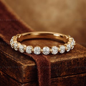 Könnte beinhalten: Ein goldener Bandring mit einer Reihe runder, klarer Edelsteine. Der Ring wird auf einer Holzbox mit einem braunen Samtband präsentiert. Das Design des Rings ist schlicht und elegant, passend für jeden Anlass.