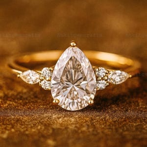 以下が含まれることがあります： 洋梨型のダイヤモンドを中央に配したゴールドの婚約指輪。メインストーンの両側に小さなアクセントダイヤモンドが施され、ゴールドのバンドにセットされています。指輪は、質感のある茶色の表面に展示されています。