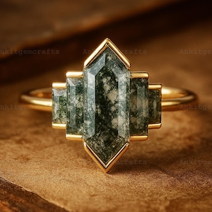 May include: A gold ring featuring a central, geometric-shaped moss agate stone with green and white inclusions. The ring has a gold band and a gold bezel setting that frames the stone. The ring is displayed on a brown surface.