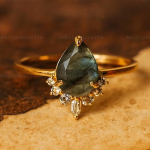 Può includere: Un anello d'oro con una pietra preziosa iridescente a forma di pera e un gruppo di piccole pietre trasparenti. La fascia e l'incastonatura dell'anello sono dorate. L'anello è posto su uno sfondo caldo e strutturato.