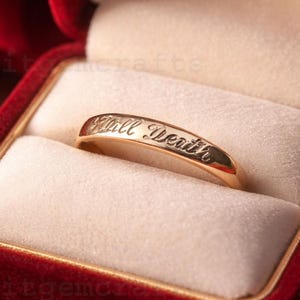 Könnte beinhalten: Ein goldener Ring mit der Inschrift "Till Death" in einer roten Samt-Ringbox. Die Ringbox ist geöffnet und zeigt den Ring vor einem cremefarbenen Innenraum. Rote Rosen sind im Hintergrund unscharf.