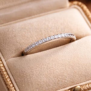 Puede incluir: Un anillo de plata con una banda de pequeñas piedras preciosas transparentes engastadas en fila. El anillo se muestra dentro de un joyero beige con ribete y cierre dorados. La caja está sobre una superficie marrón.