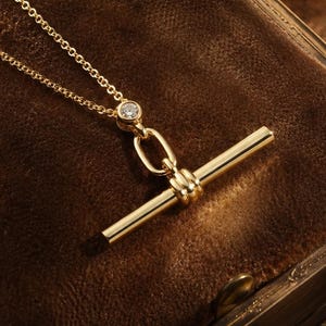 Könnte beinhalten: Eine zarte goldene Halskette mit einem Stabanhänger und einem kleinen Diamantakzent. Die Halskette wird auf einer braunen, strukturierten Oberfläche präsentiert, was eine Vintage-Ästhetik erzeugt. Die Kette ist fein und der Anhänger ist zentriert.