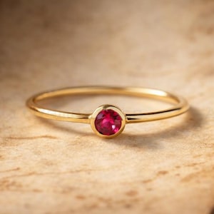 May include: A delicate gold ring featuring a vibrant, round ruby gemstone. The ring has a thin, polished band and a bezel setting that securely holds the red gemstone. The ring is displayed on a textured, neutral-toned surface.