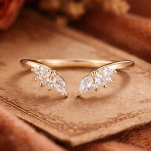 Könnte beinhalten: Ein goldfarbener Ring mit geteiltem Schaft-Design. Jede Seite des Rings weist eine Ansammlung von marquiseförmigen und runden, klaren Edelsteinen auf, die ein zartes, elegantes Aussehen erzeugt. Der Ring ist vor einem weichen, warm getönten Hintergrund platziert.