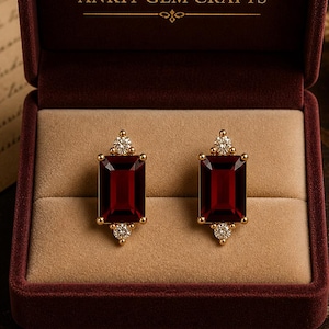 Può includere: Un paio di orecchini in oro con pietre preziose rettangolari rosso scuro e piccoli accenti di diamanti, presentati in un portagioie di velluto bordeaux aperto. Il nome del marchio "ANKIT GEM CRAFTS" è visibile.
