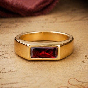 May include: A gold-colored ring with a rectangular, deep red gemstone. The ring has a simple, classic design with a polished finish. The gemstone is set flush within the ring's band, creating a sleek, modern look. Jewelry.