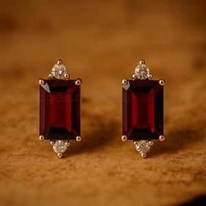 Pode incluir: Um par de brincos de botão dourados. Cada brinco apresenta uma pedra preciosa retangular vermelha escura, acentuada com pedras preciosas claras menores acima e abaixo. Os brincos têm um design clássico e elegante.