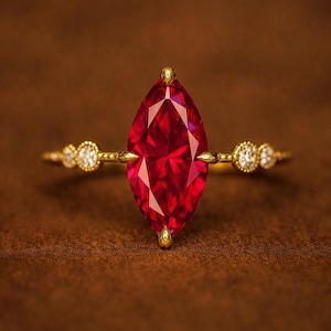 May include: A gold ring with a striking, marquise-cut, deep red gemstone. The band is slender, with small, round, clear gemstones on either side of the main stone. The ring is set against a warm brown backdrop.