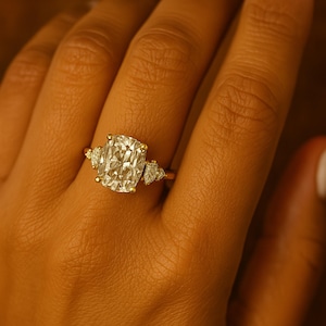 May include: A gold ring featuring a large, cushion-cut diamond as the centerpiece, flanked by smaller, pear-shaped diamonds on each side. The ring is set on a gold band, showcasing a classic and elegant design.