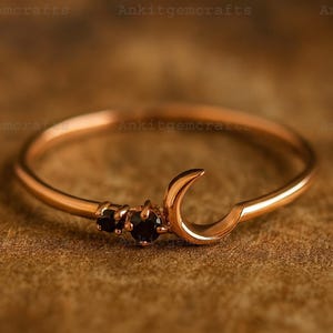 May include: A rose gold ring featuring a crescent moon design with two small black gemstones. The ring has a delicate band and a polished finish, set against a warm brown background.