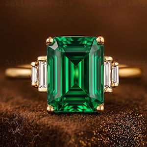 May include: A gold ring with a large, emerald-cut green gemstone at its centre, with smaller, rectangular white gemstones on either side. The ring has a gold band and gold accents. The text "Ankitgemcrafts" is visible.