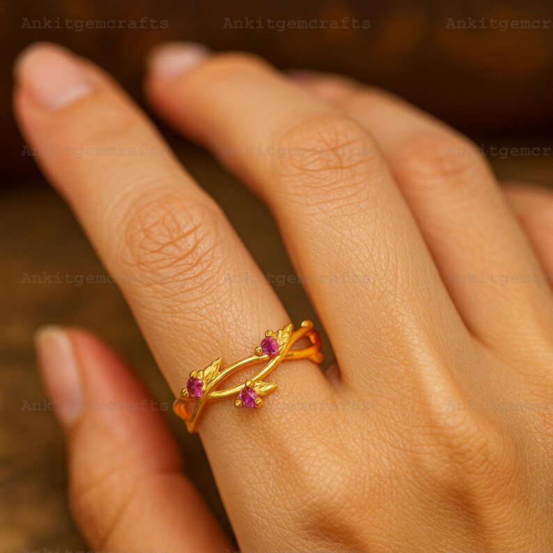 May include: A gold-colored ring with a leaf design and three small, round, pink gemstones. The ring is on a finger, and the design wraps around the finger. The ring is likely made of gold-plated metal.