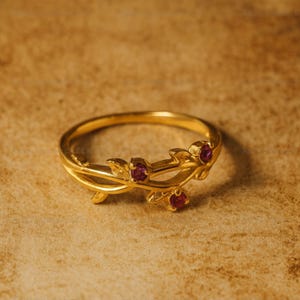 May include: A gold-colored ring with a delicate vine design. The ring features small, round, red gemstones set along the vine. The ring is displayed on a textured, tan surface. Jewelry.