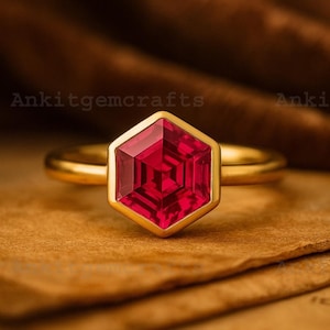 May include: A gold ring featuring a hexagonal, faceted, deep red gemstone. The ring has a simple, polished gold band. The gemstone is set in a gold bezel. The background is a warm, textured brown.