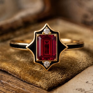 May include: A gold ring with a rectangular, deep red gemstone at the center, framed by a black enamel design. Two small diamonds are set on the black enamel. The band has a black enamel stripe. The ring is displayed on a textured, golden surface.