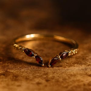 Può includere: Un anello dorato con un design a forma di V. L'anello presenta diverse pietre preziose rosso scuro taglio marquise. La fascia è sottile e delicata, con piccoli accenti rotondi. L'anello è posto su uno sfondo marrone caldo.